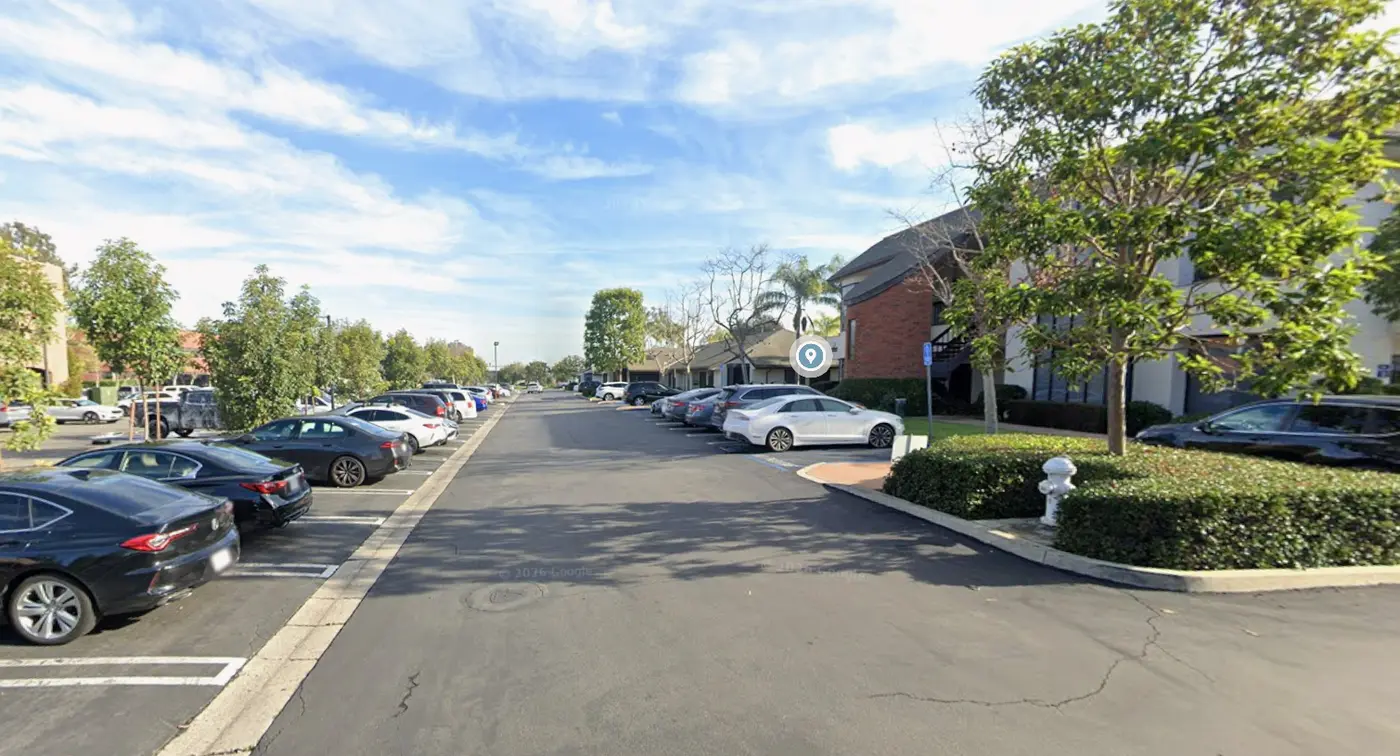 Street-level view approaching the Costa Mesa business campus, with parked cars lining the drive and the two-story brick-and-stucco office buildings ahead on the right